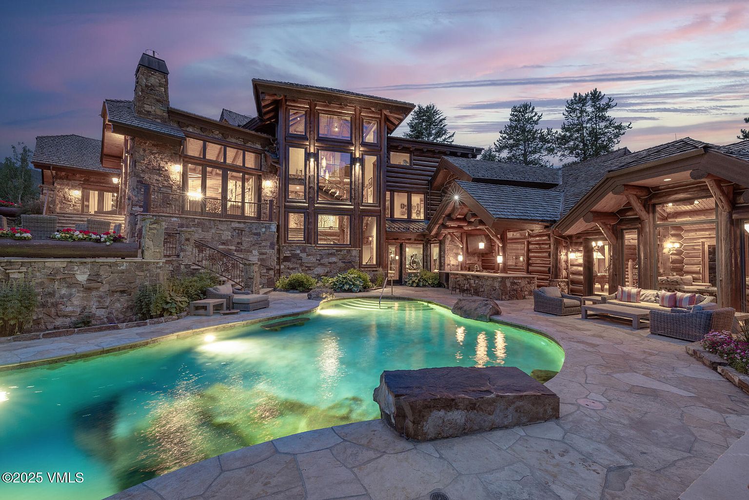This image showcases the rear exterior of a luxurious log cabin home, highlighting a stunning pool and spa area. The pool features a unique shape and is illuminated with a vibrant turquoise glow, complemented by a stone patio with comfortable seating. The architecture combines stone and wood elements, creating a rustic yet elegant ambiance under a twilight sky.
