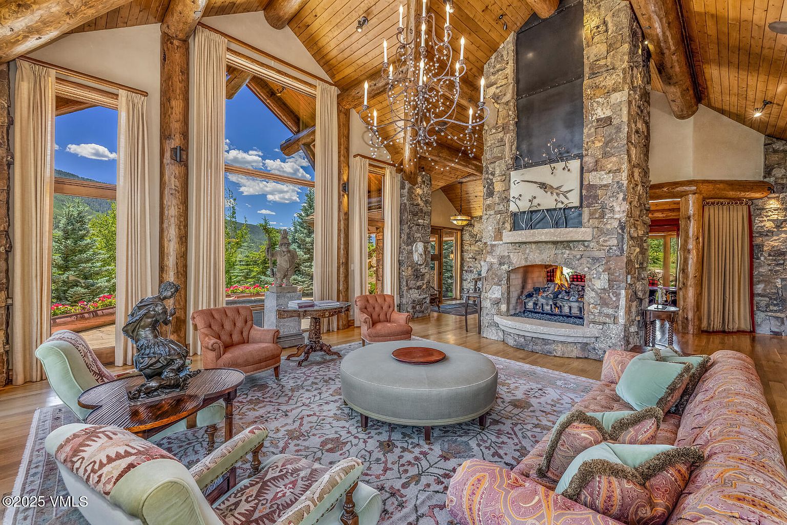 This grand living room features a soaring wood-paneled ceiling, a massive stone fireplace, and large windows offering scenic views. The room is furnished with comfortable seating arranged around a central ottoman, all set upon an ornate area rug. The overall impression is one of rustic luxury and spaciousness, perfect for entertaining or relaxing.