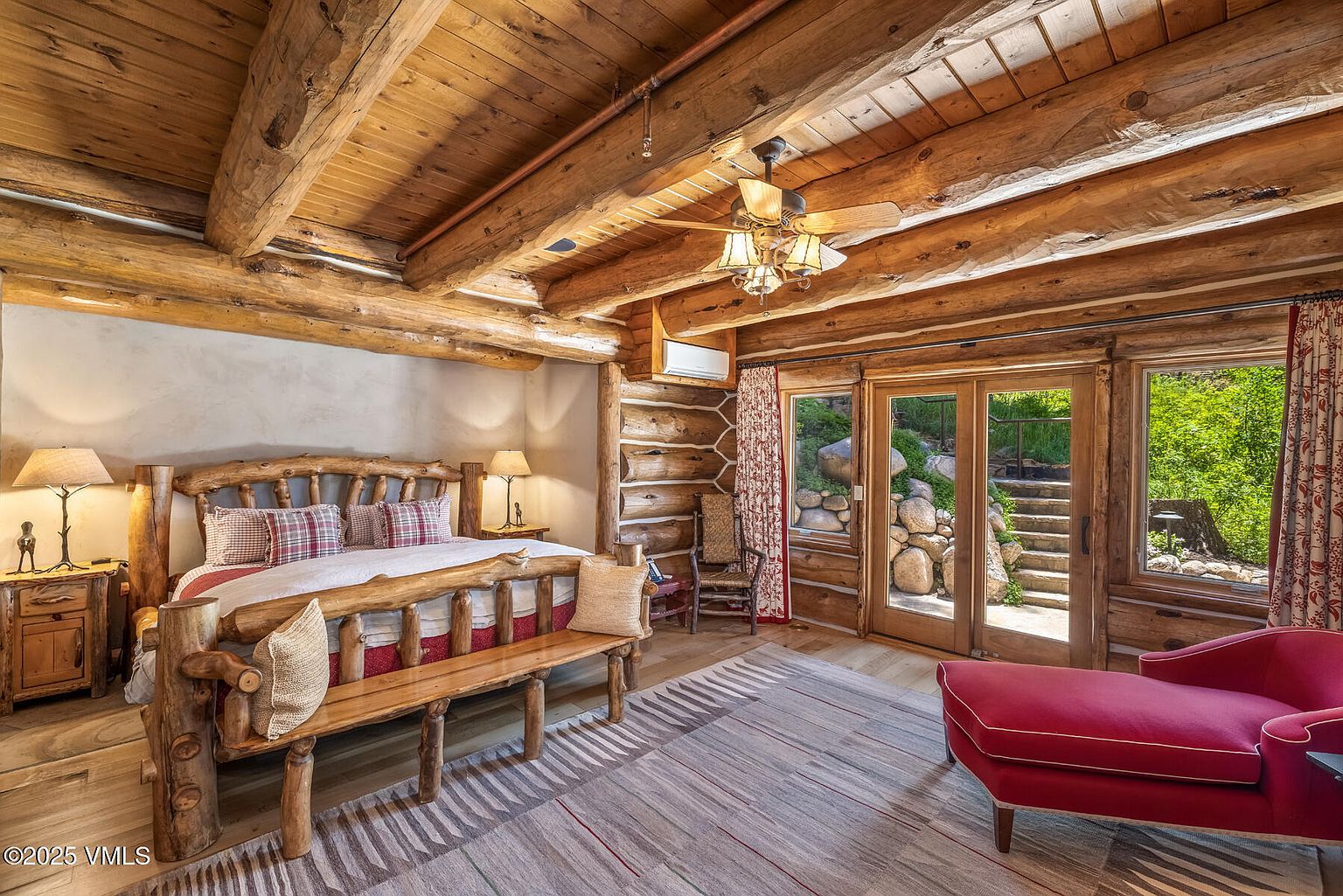 This is a cozy primary bedroom featuring a rustic log cabin design with exposed wooden beams and a log bed frame. The room is well-lit with natural light streaming through the windows and glass doors, which lead to an outdoor area. A red chaise lounge adds a pop of color, and a patterned rug ties the space together, creating a warm and inviting atmosphere.