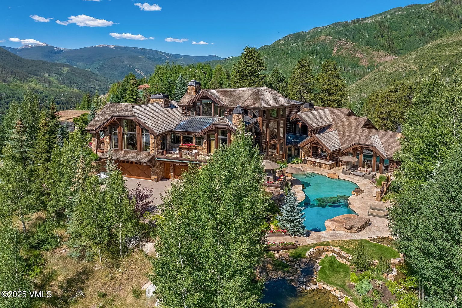 This aerial view showcases a luxurious mountain home with a sprawling layout, featuring a combination of stone and wood construction. The property includes a large pool with a stone surround, a pond, and is surrounded by lush greenery and mountain views. The architectural style is rustic and elegant, creating a sense of privacy and exclusivity.