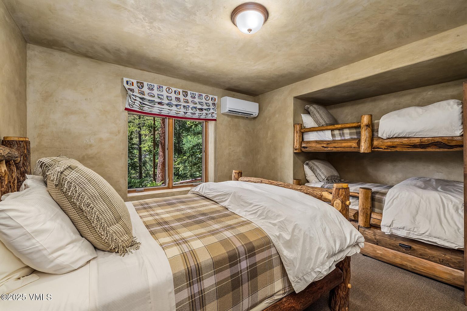 This is a cozy bedroom featuring a log-style bed and built-in bunk beds, creating a rustic and inviting atmosphere. The room is decorated with a plaid bedspread and neutral-toned pillows, complemented by a window offering a view of the surrounding trees. The walls have a textured finish, adding to the room's warm and natural aesthetic.