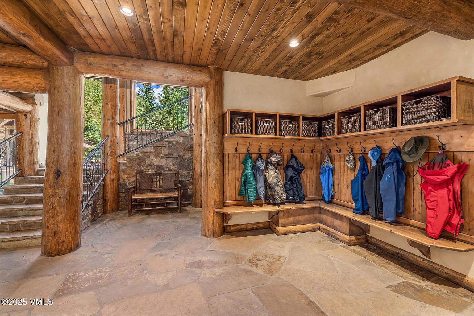 This interior shot showcases a rustic hallway and staircase area. The space features natural stone flooring, exposed wooden beams and pillars, and a built-in mudroom area with wooden benches, hooks, and storage baskets. A stone staircase with wrought iron railings leads to another level, creating a warm and inviting atmosphere.