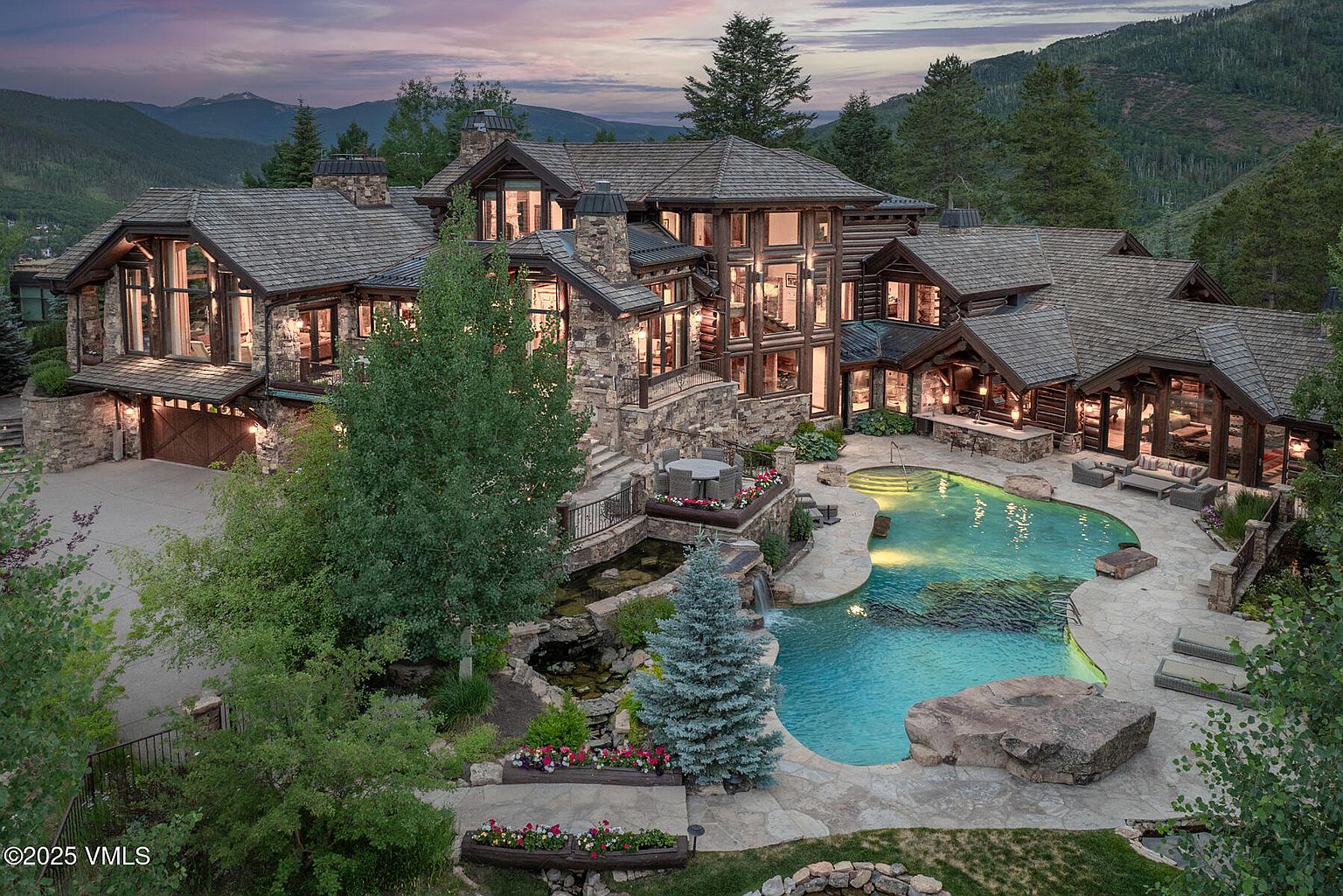 This aerial view showcases a luxurious mountain estate with a stunning stone and timber design. The property features a large pool with a waterfall, meticulously landscaped gardens, and multiple outdoor living spaces. The architecture blends seamlessly with the natural surroundings, creating a sense of rustic elegance and privacy.