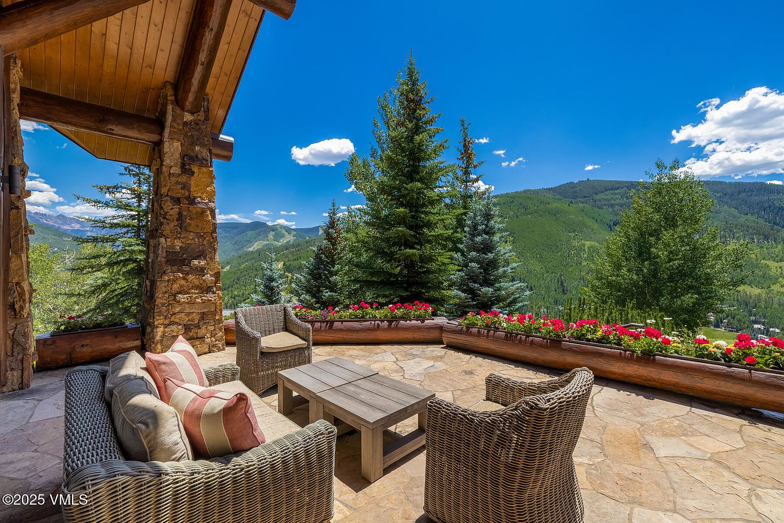 This image showcases a luxurious outdoor patio area with comfortable wicker furniture, including a sofa and chairs, arranged around a wooden coffee table. The patio is constructed with natural stone and features built-in planters filled with vibrant red and white flowers. The backdrop offers a stunning view of lush green mountains under a clear blue sky, creating a serene and inviting atmosphere.