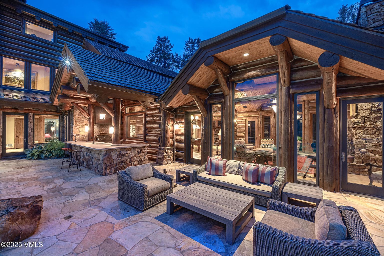 This image showcases a luxurious outdoor patio area of a log cabin home. The patio features comfortable wicker furniture, including a sofa, armchairs, and a coffee table, arranged on a stone-paved surface. The log cabin structure is visible in the background, with large windows offering views into the interior, and a stone bar area is also present, suggesting an ideal space for relaxation and entertaining.