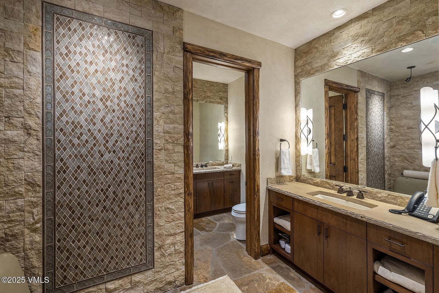 This is a well-appointed primary bathroom featuring stone walls and flooring, creating a rustic yet luxurious feel. The vanity has dark wood cabinetry and a light countertop, complemented by a large mirror and decorative sconces. An adjacent bathroom area is visible through a wooden-framed doorway, adding depth to the space.