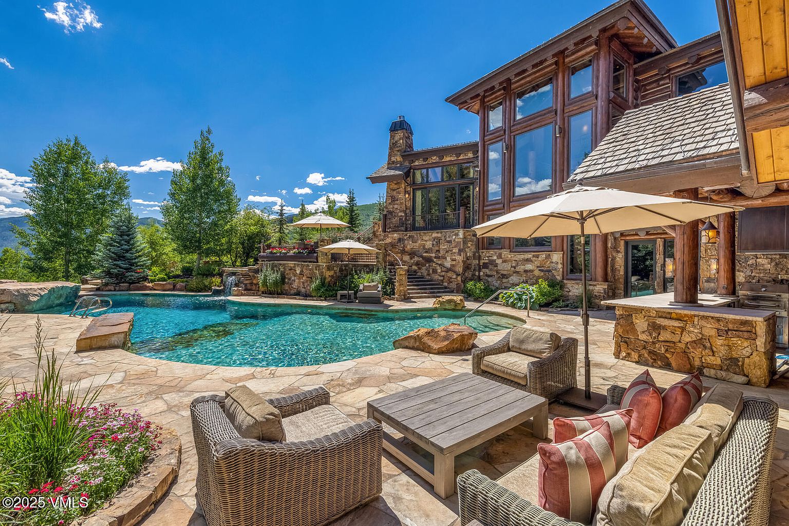 This image showcases a luxurious outdoor pool and patio area of a high-end home. The pool features clear blue water and a natural stone surround, complemented by lush landscaping. The patio includes comfortable wicker furniture, a stone-clad outdoor bar area, and an umbrella, creating an inviting space for relaxation and entertainment. The house is built with stone and wood, with large windows that reflect the sky.
