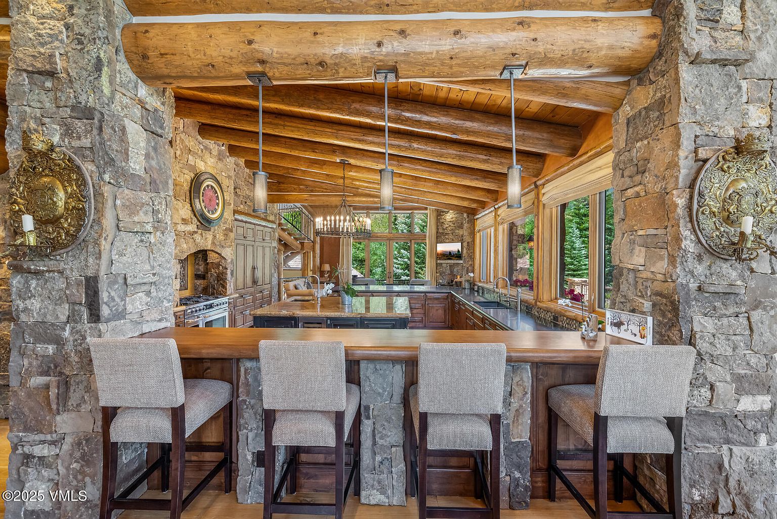 This is a view of a rustic kitchen featuring stone pillars and a wooden beam ceiling. The kitchen has a breakfast bar with four chairs, dark wood cabinetry, and stainless steel appliances. The space is well-lit with pendant lighting and natural light from the windows, creating a warm and inviting atmosphere.
