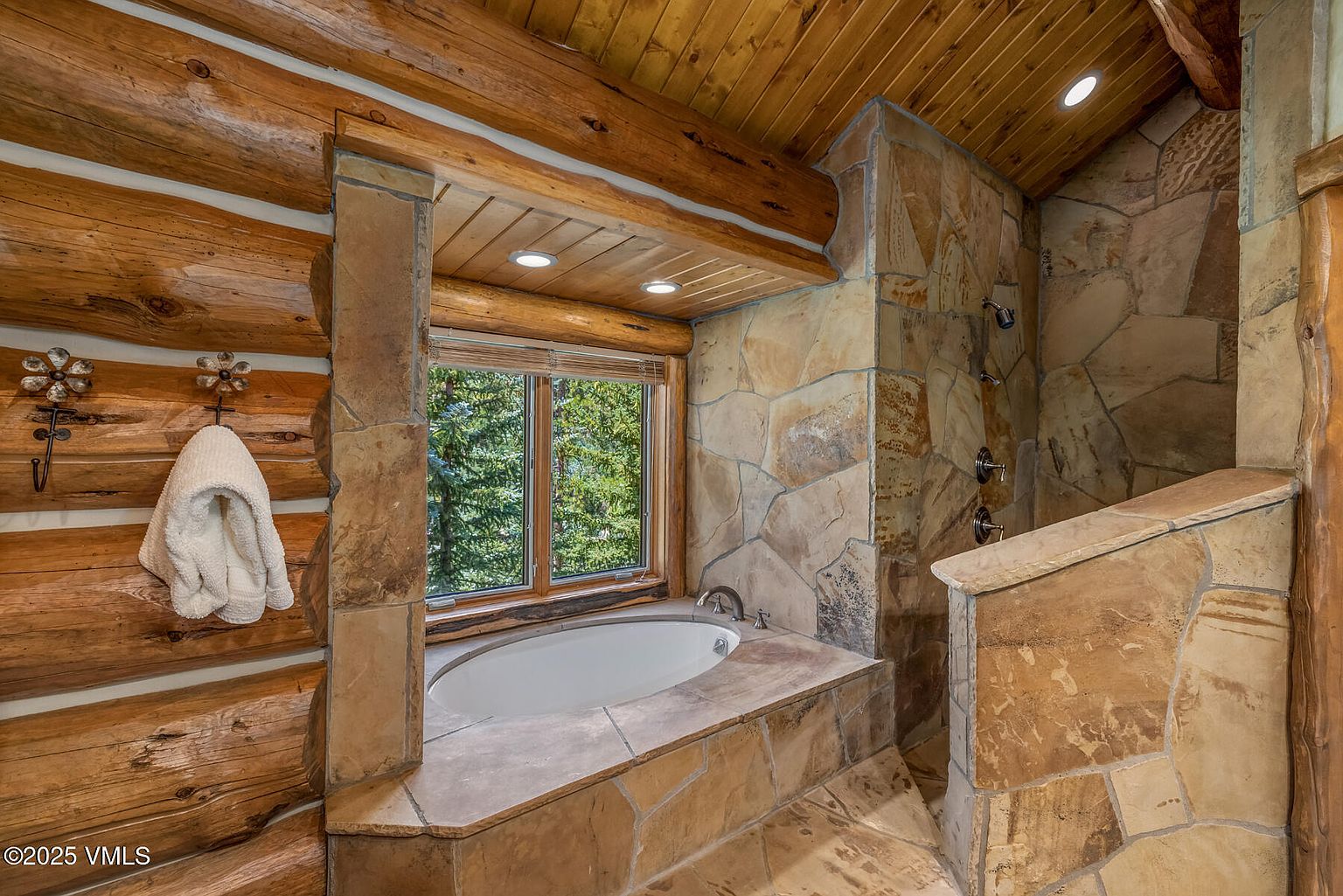 This is a luxurious primary bathroom featuring a rustic design with log walls and stone accents. A built-in bathtub sits beneath a window offering a view of the outdoors, while a stone-clad shower is located to the right. The bathroom exudes a spa-like atmosphere with its natural materials and warm lighting.