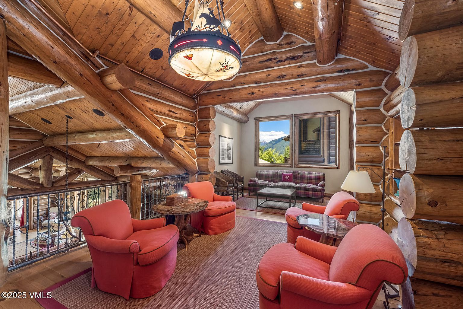 This is an interior shot of a rustic living room featuring log walls and ceiling beams, creating a warm and inviting atmosphere. The room is furnished with several red armchairs, wooden tables, and a plaid sofa visible through an opening. A unique chandelier hangs from the ceiling, adding to the room's character and charm.