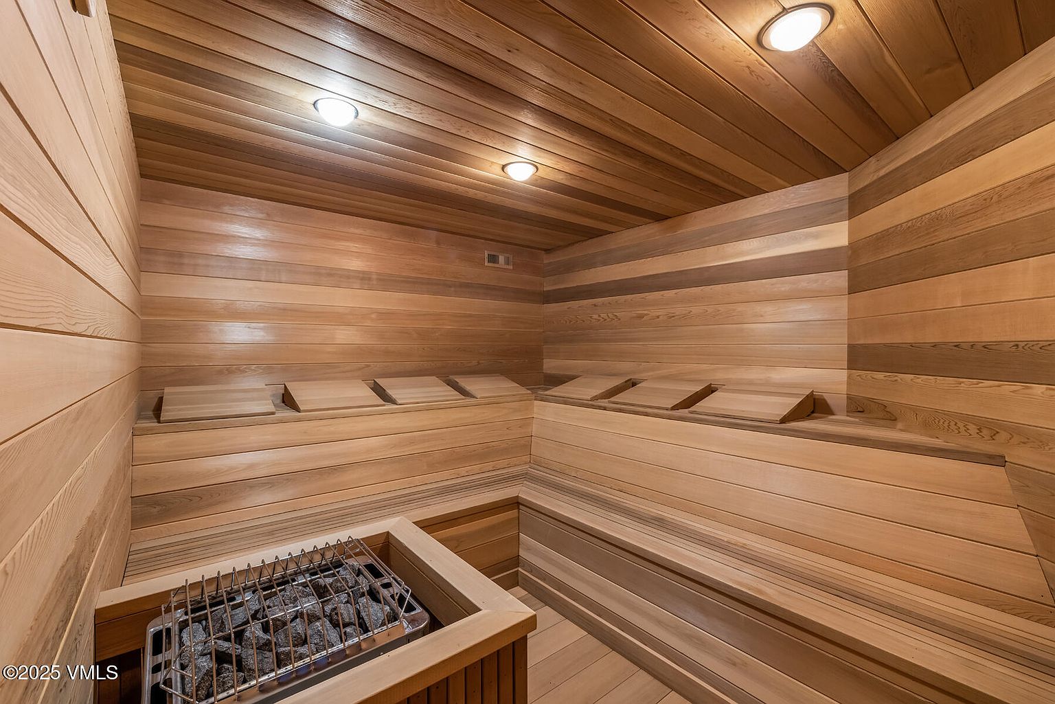 This is an interior shot of a sauna, featuring wooden walls, ceiling, and benches. A heater with rocks sits in the foreground, and recessed lighting illuminates the space. The sauna appears clean and well-maintained, offering a relaxing and luxurious amenity.