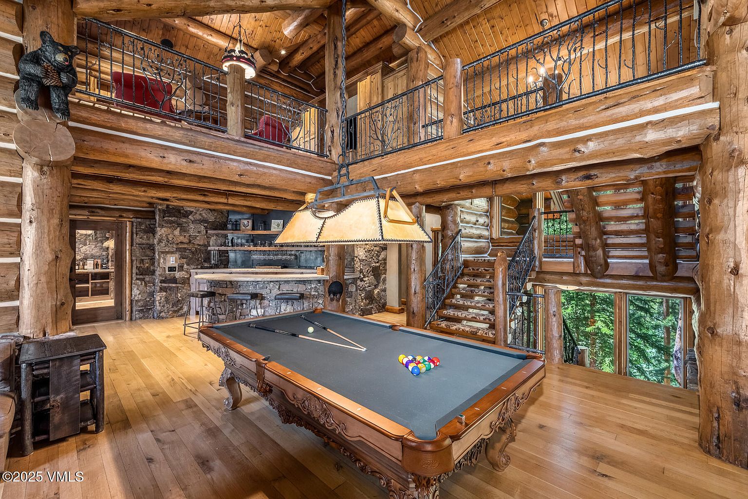 This grand living room features a rustic log cabin design with exposed wooden beams and a stone accent wall. A pool table takes center stage, illuminated by a unique overhead light fixture. The room also includes a bar area and a staircase leading to an upper level, creating a spacious and inviting atmosphere.