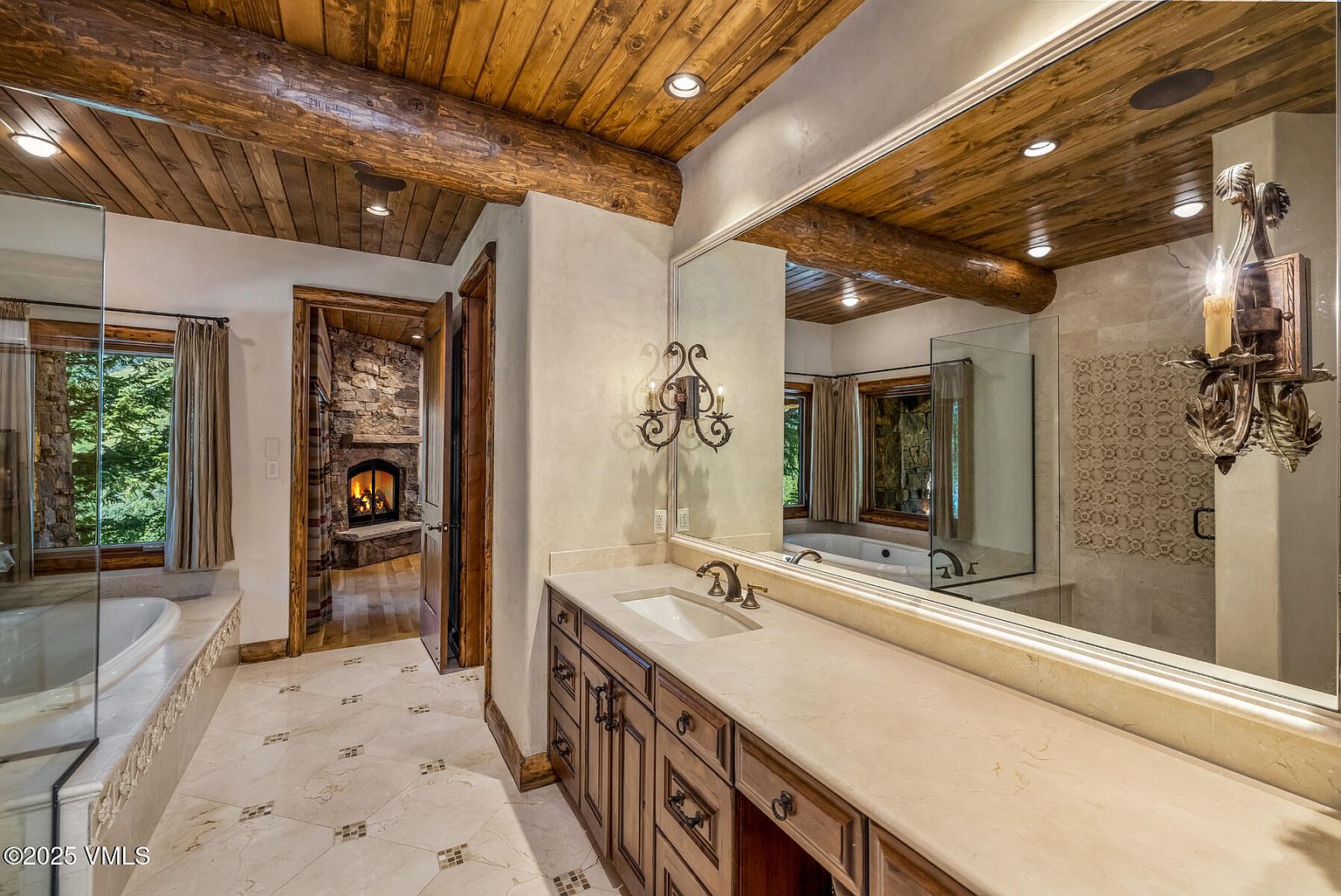 This is a luxurious primary bathroom featuring a wooden beamed ceiling and a large vanity with a light-colored countertop and dark wood cabinetry. A large mirror reflects the space, including a glimpse of a soaking tub and a stone fireplace in an adjacent room. The flooring is a light tile with decorative mosaic accents, contributing to the bathroom's elegant and rustic-chic aesthetic.