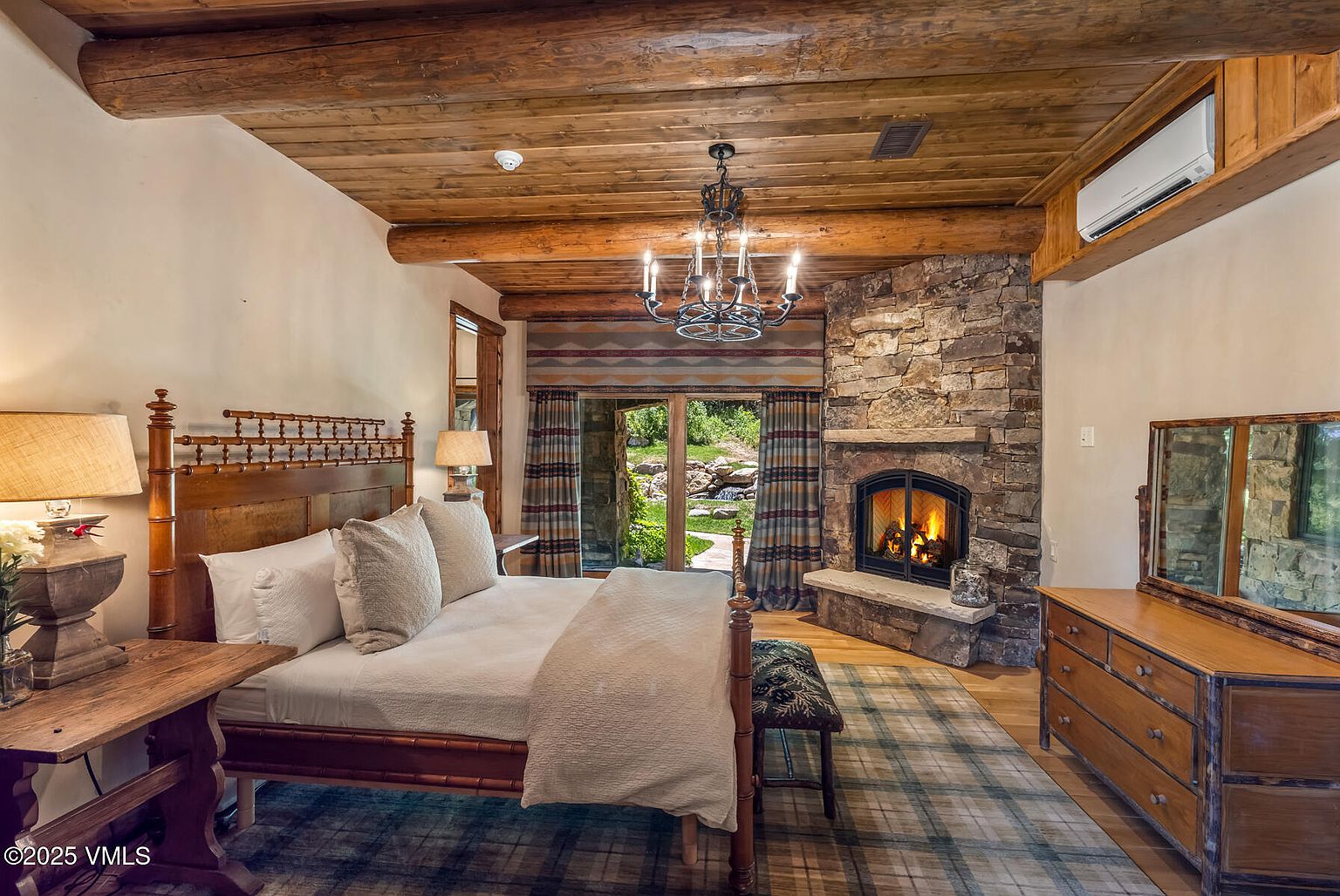 This is a warm and inviting primary bedroom featuring a wooden four-poster bed with plush white bedding, a stone fireplace, and exposed wooden beams on the ceiling. The room is well-lit by a chandelier and lamps, creating a cozy and luxurious atmosphere. A plaid rug adds a touch of rustic charm, complementing the natural materials used throughout the space.