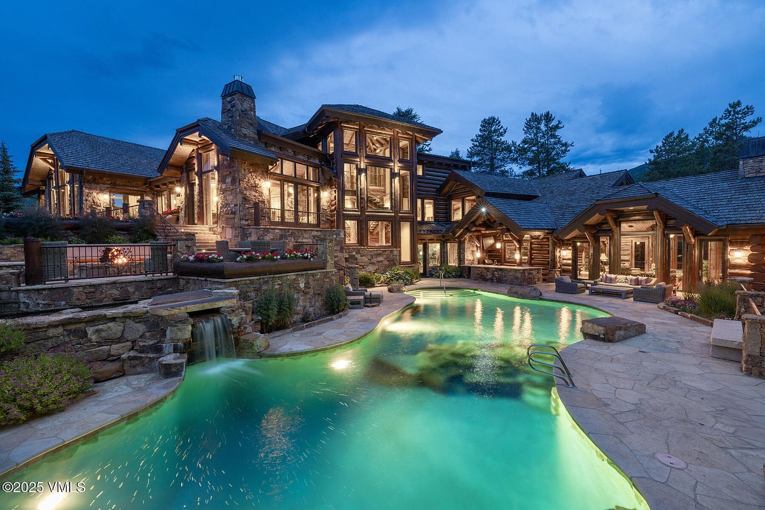 This image showcases the rear exterior of a luxurious log cabin home, highlighting a stunning pool and spa area. The pool features a naturalistic design with integrated waterfall and stone accents, illuminated by underwater lighting. The home itself is constructed with a combination of stone and log elements, exuding rustic elegance and creating an inviting outdoor living space.