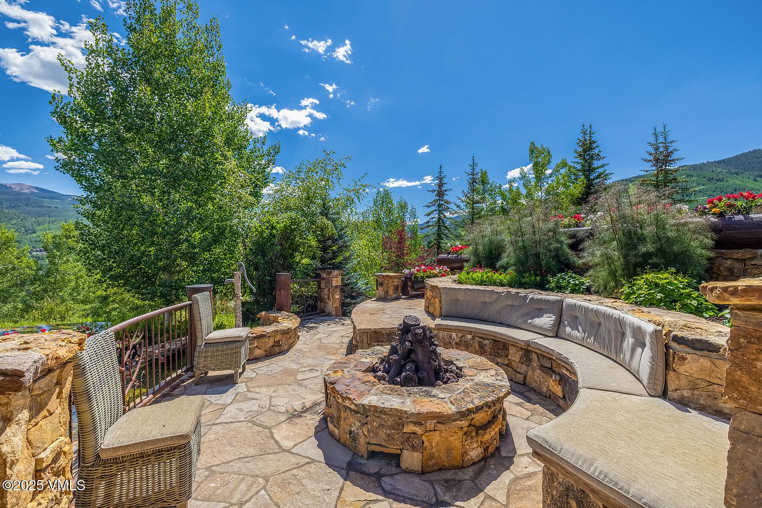 This image showcases a beautifully designed outdoor patio area, featuring a stone fire pit surrounded by a curved stone bench with comfortable cushions. The patio is constructed with natural stone pavers and is bordered by lush landscaping, including trees, shrubs, and flowering plants. The scene evokes a sense of relaxation and outdoor living, perfect for entertaining or enjoying quiet moments.