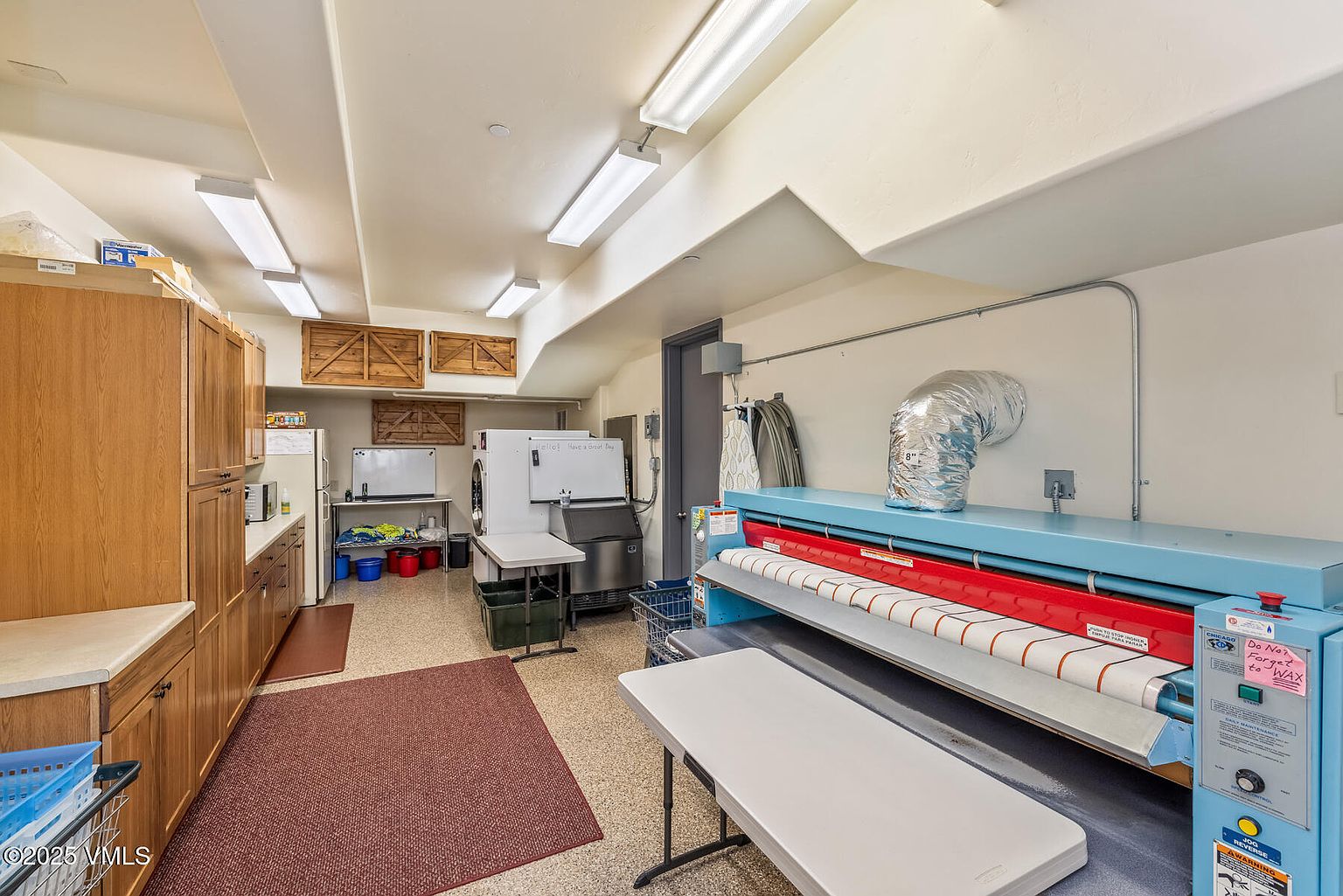 This is an interior shot of a commercial laundry room. The room features a large commercial ironing machine, a washer and dryer, and ample cabinet space. The flooring is speckled, and the walls are painted in a neutral tone. The room is well-lit with fluorescent lighting.