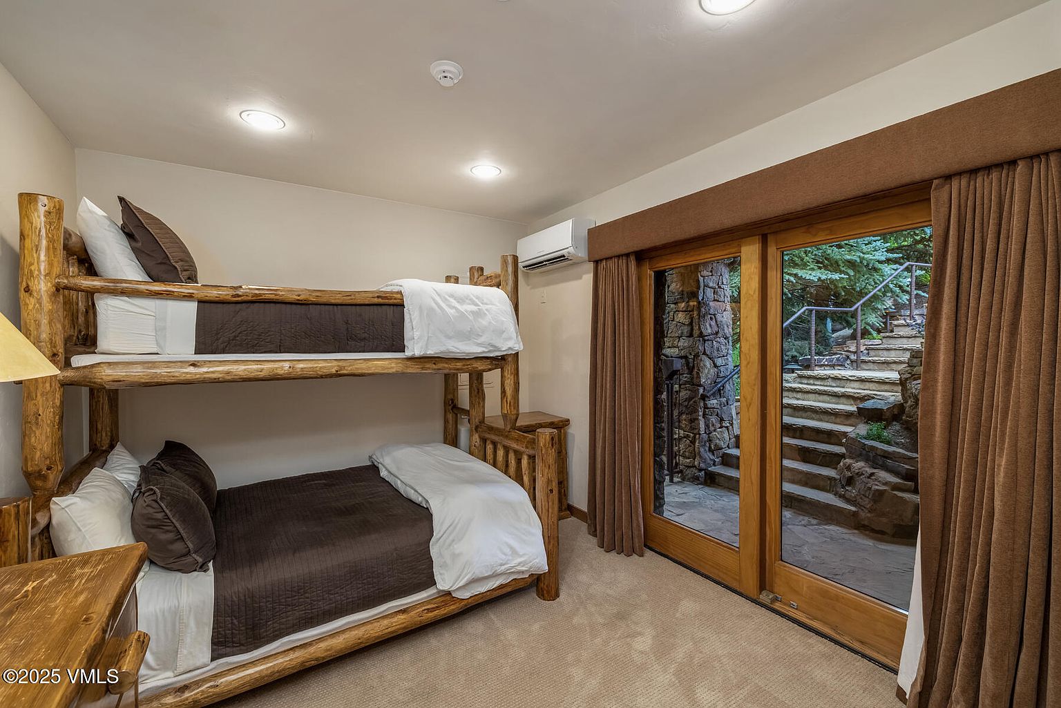 This is a cozy guest bedroom featuring a rustic log bunk bed with brown bedding and white linens. A sliding glass door leads to an outdoor stone staircase, providing natural light and a unique view. The room is carpeted and has a warm, inviting atmosphere.