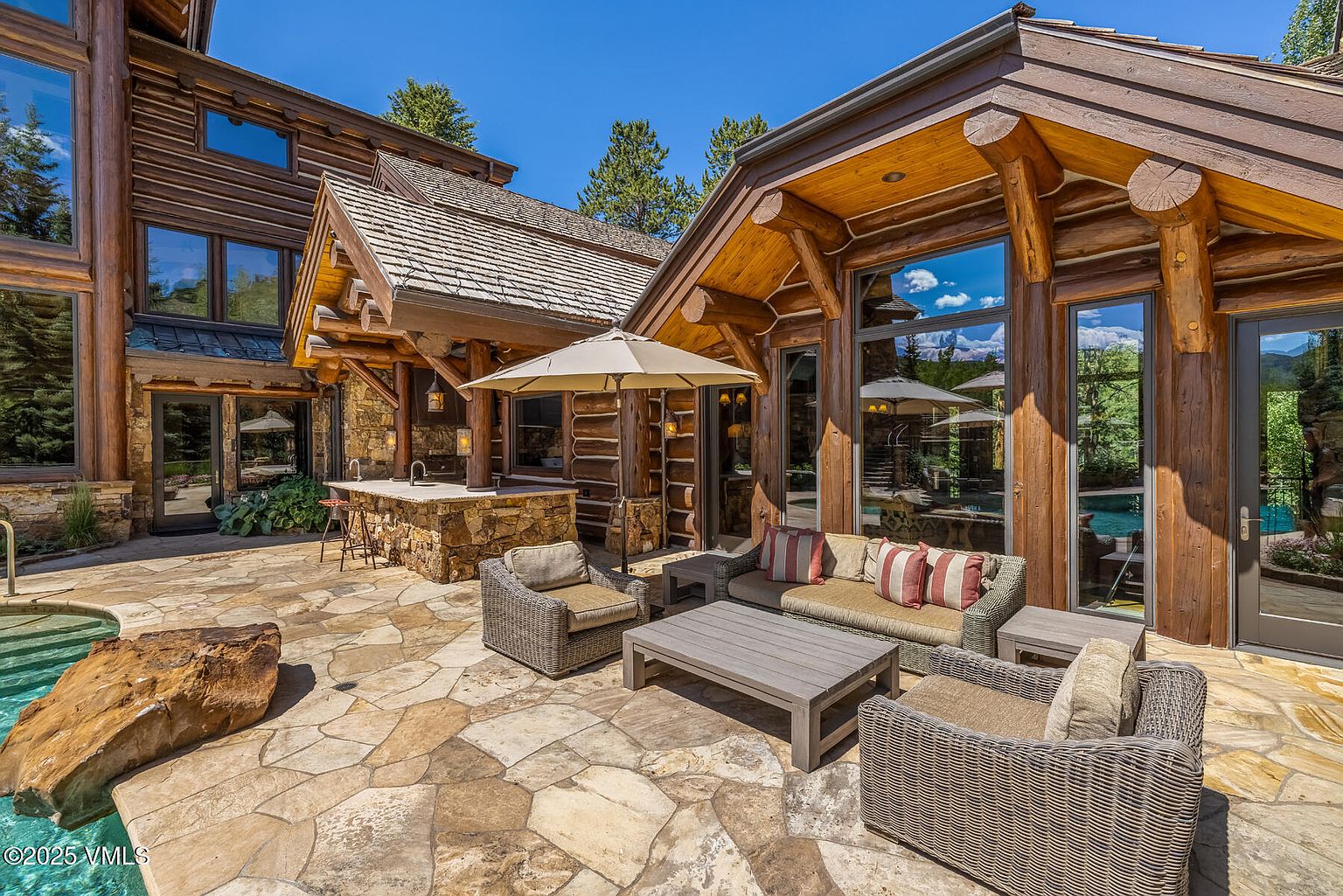 This image showcases a luxurious outdoor patio area, featuring stone flooring, comfortable wicker furniture, and a built-in stone bar. The patio is adjacent to a log cabin-style home with large windows that reflect the surrounding landscape. The scene exudes a sense of relaxation and upscale outdoor living.