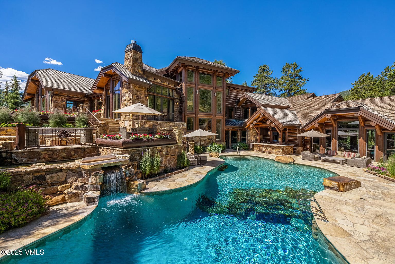 This image showcases the rear exterior of a luxurious log cabin home, emphasizing a stunning pool and spa area. The pool features a naturalistic design with integrated rock features and a small waterfall, surrounded by a stone patio. The home itself is constructed with a combination of wood and stone, featuring large windows that offer views of the surrounding landscape, creating an inviting and upscale outdoor living space.