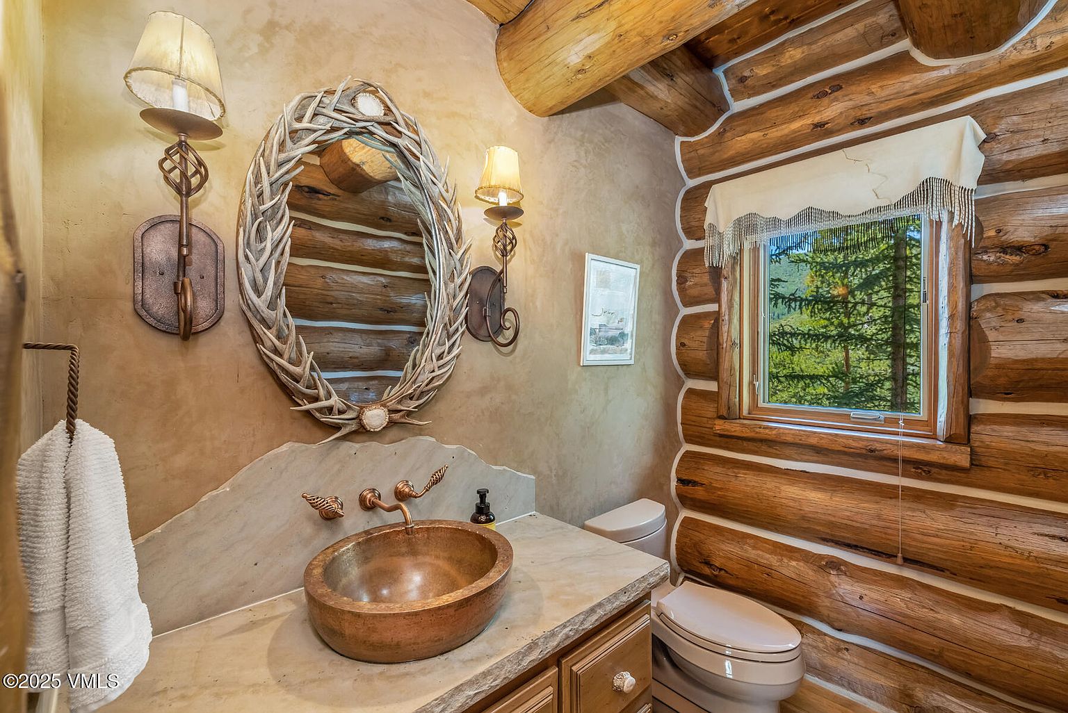 This is a rustic bathroom featuring log walls and a unique antler-framed mirror. The copper sink and stone countertop add to the natural aesthetic. A small window provides natural light and a view of the outdoors, enhancing the cabin-like feel.