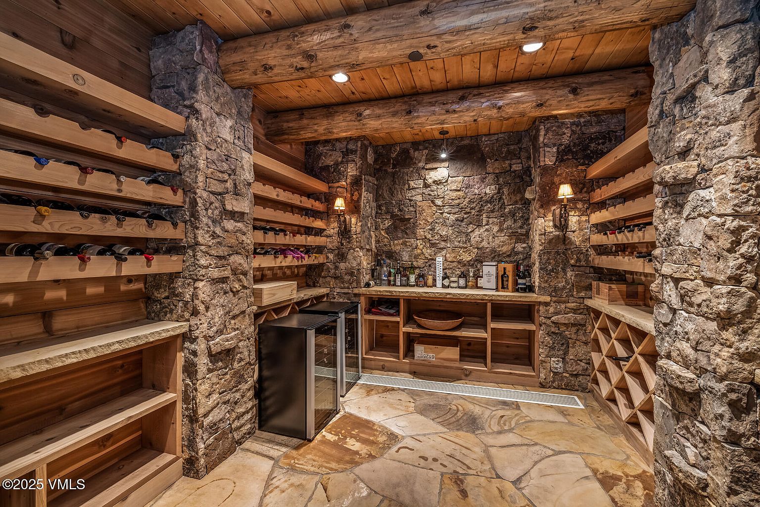 This is a rustic wine cellar featuring stone walls and wooden shelving filled with wine bottles. The floor is made of stone tiles, and the ceiling has exposed wooden beams. A small wine refrigerator is present, adding a modern touch to the otherwise traditional design.