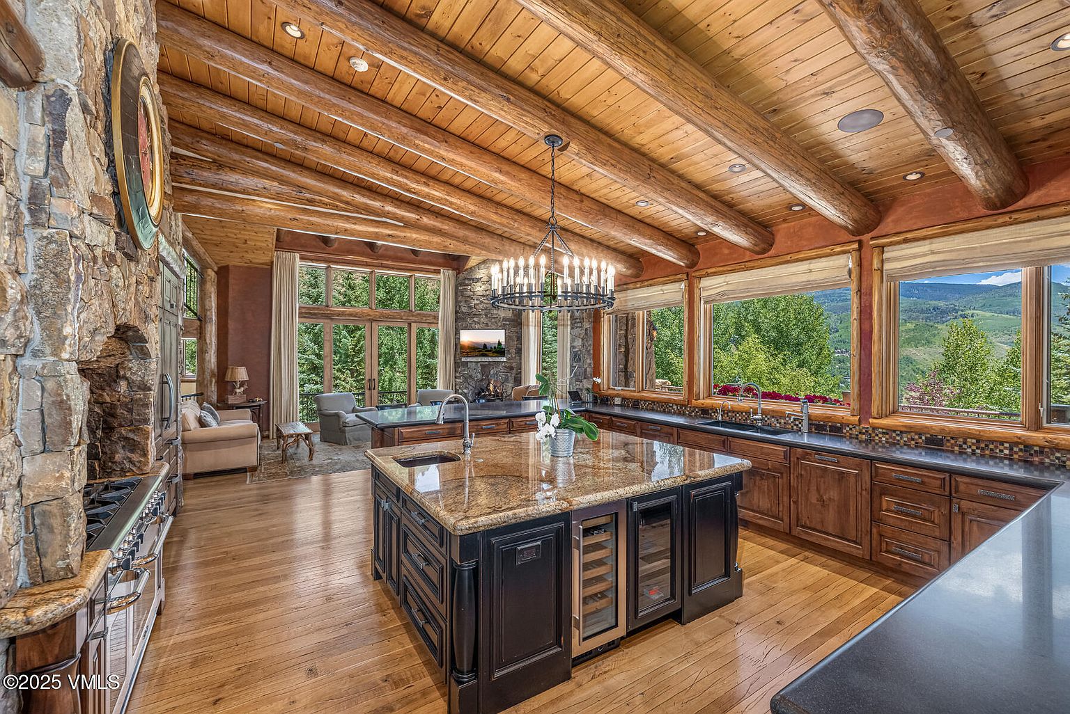 This is a spacious, rustic kitchen featuring a large central island with a granite countertop and a built-in wine cooler. The kitchen boasts wooden cabinetry, a stone accent wall, and a high ceiling with exposed wooden beams. Large windows offer scenic views of the surrounding landscape, creating a warm and inviting atmosphere.