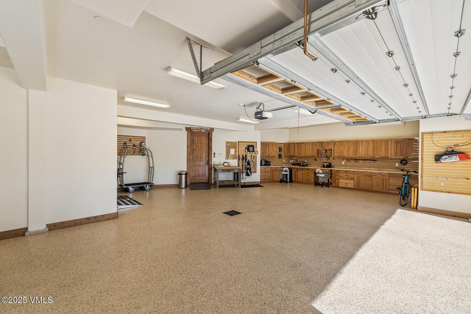 This is a spacious and well-organized garage. It features custom wooden cabinetry along one wall, providing ample storage and workspace. The floor is finished with a durable epoxy coating, and the garage door is a modern, insulated design. The overall impression is clean, functional, and high-end.