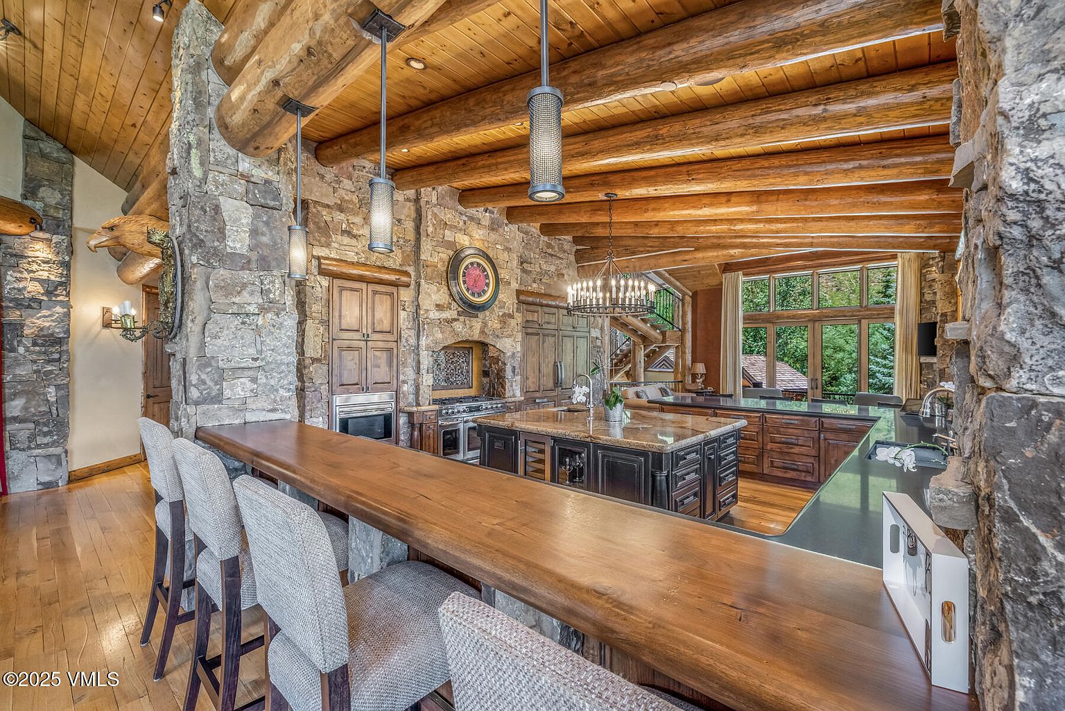 This is a rustic kitchen featuring a stone wall, wooden beams, and hardwood floors. The kitchen island has a granite countertop and dark wood cabinetry. There is a long wooden bar with upholstered stools, and the kitchen is well-lit with pendant lights and a chandelier.
