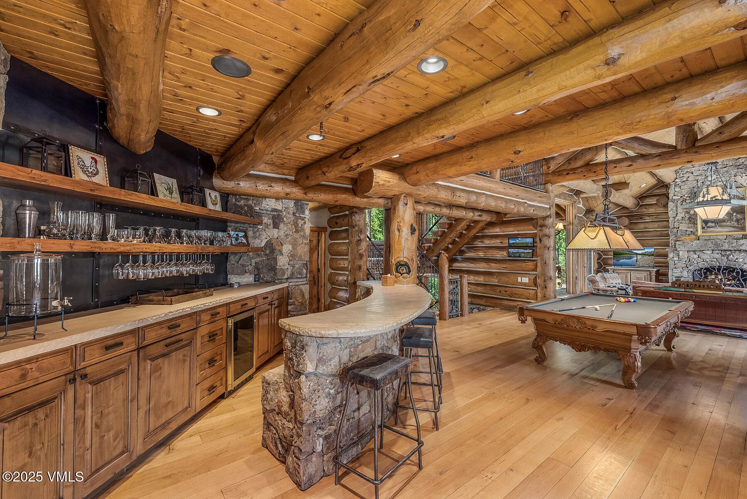 This interior shot showcases a rustic living room with a log cabin aesthetic. The room features exposed wooden beams, a stone fireplace, a bar area with wooden cabinets and stone accents, and a pool table. The hardwood floors and warm lighting create a cozy and inviting atmosphere.