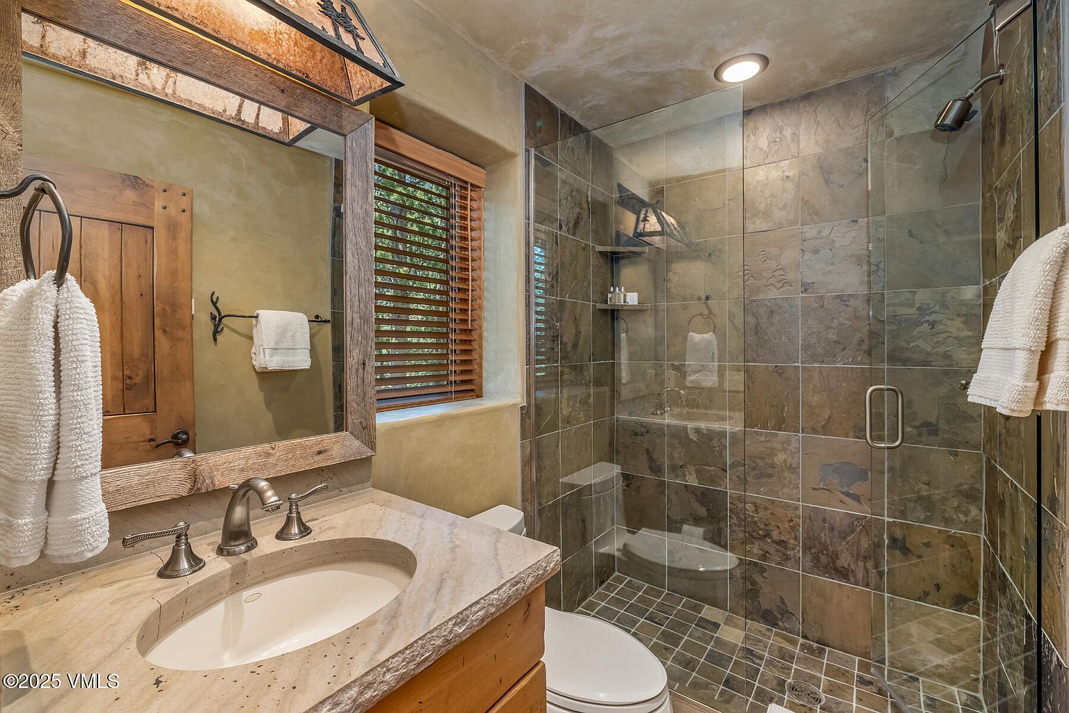 This is a bathroom featuring a rustic design with a stone countertop vanity, wooden cabinetry, and a framed mirror. The shower is enclosed with glass and has tiled walls, while the floor is also tiled. The overall impression is a cozy and well-maintained space.