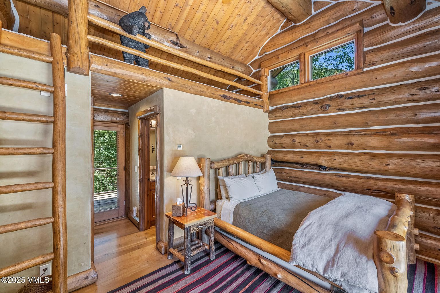 This is a cozy bedroom with a rustic log cabin aesthetic. The walls are constructed from exposed logs, complemented by a wooden ceiling and a decorative bear statue on a high shelf. A wooden bed frame and matching nightstand enhance the room's natural charm, while a ladder leads to an upper level, and a doorway provides access to an outdoor balcony.
