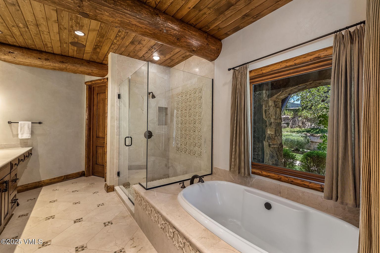 This is a primary bathroom featuring a glass-enclosed shower, a freestanding bathtub, and a window with a view of the outdoors. The bathroom has a rustic-chic style with a wooden ceiling and beams, and the floor is tiled with a decorative pattern. The overall impression is luxurious and relaxing.