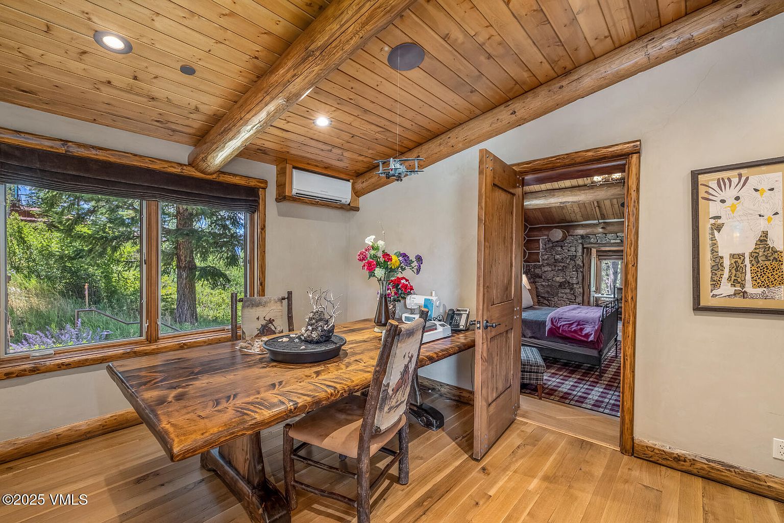 This interior shot showcases a rustic dining room with a large wooden table and chairs. The room features a wooden ceiling with exposed beams, a window with a view of greenery, and an open doorway leading to another room. The style is warm and inviting, emphasizing natural materials and a cozy atmosphere.