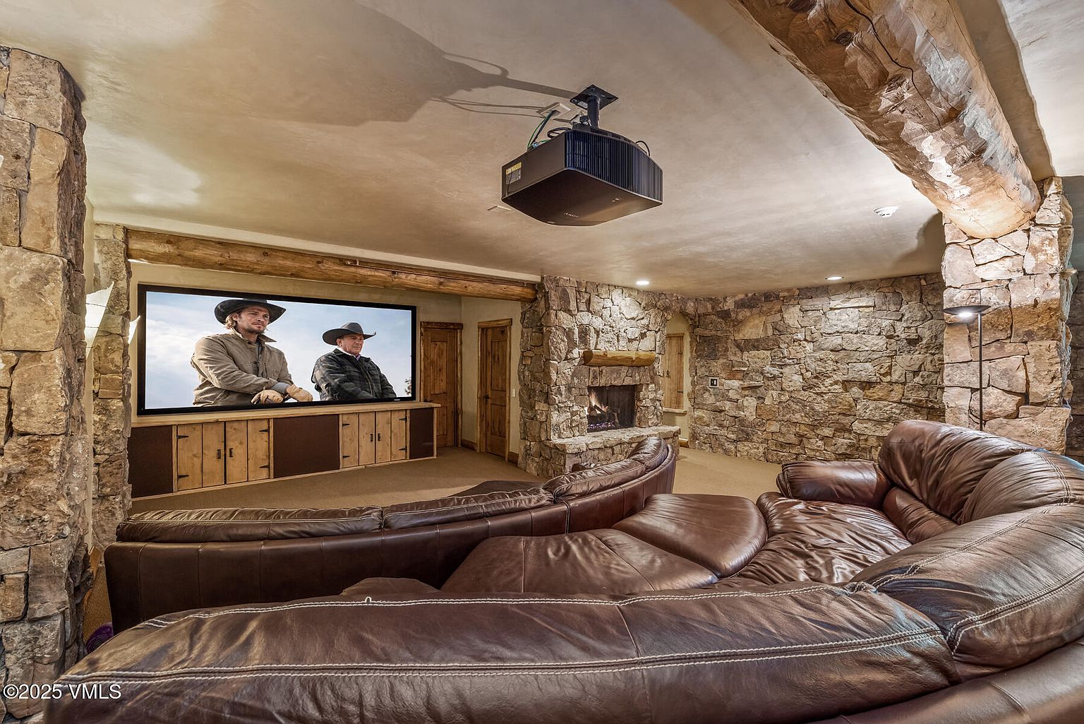 This is an interior shot of a rustic-style living room or home theater. The room features stone walls, a stone fireplace, and a large curved leather sofa. A projector displays a movie on a screen, creating a cozy and immersive entertainment space.