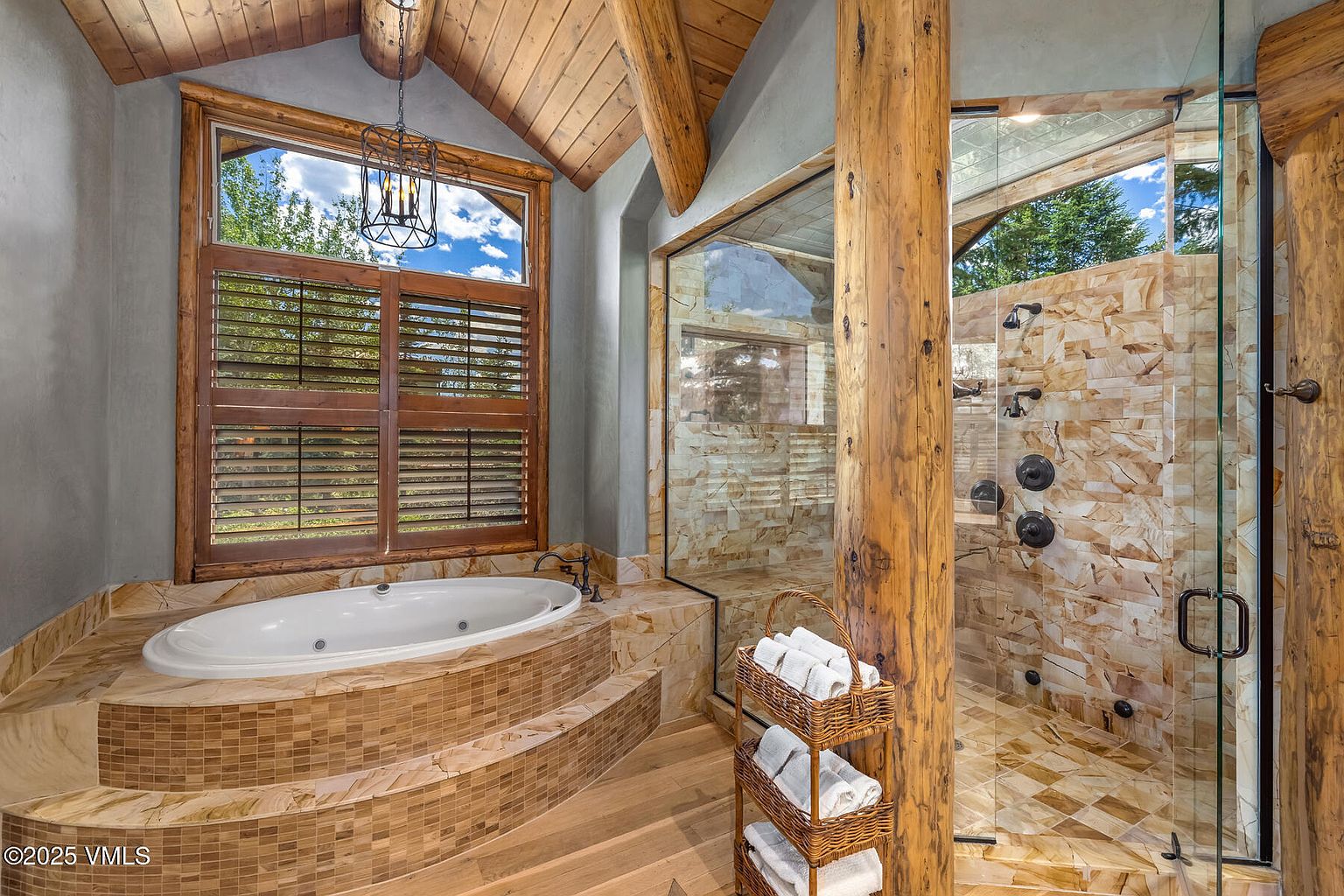 This is a luxurious primary bathroom featuring a jacuzzi tub surrounded by mosaic tile steps, a spacious glass-enclosed shower with natural stone walls, and rustic wooden support beams. The room is well-lit by a large window with wooden shutters, creating a spa-like atmosphere with a blend of natural and modern elements. A towel rack with neatly folded white towels adds to the sense of comfort and elegance.