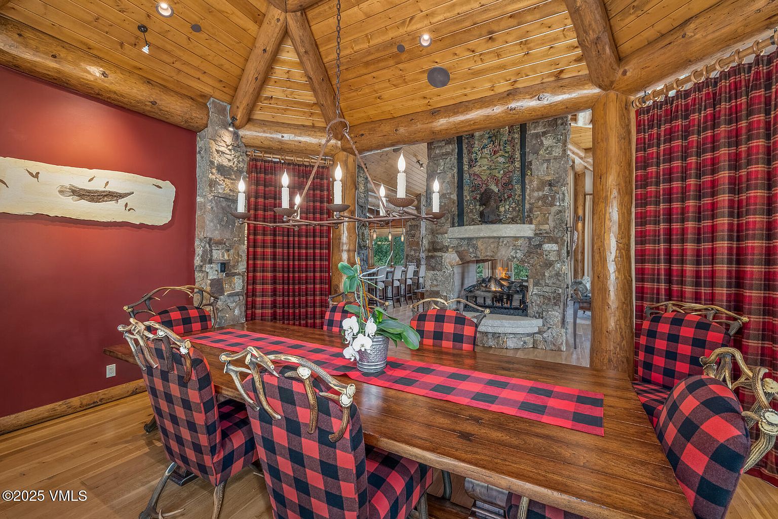 This is an inviting dining room featuring a rustic wooden table with a red and black plaid runner, surrounded by matching plaid chairs with antler-inspired details. The room boasts a warm ambiance with a stone fireplace, wooden ceiling beams, and plaid curtains, creating a cozy and luxurious atmosphere. A unique chandelier hangs above the table, adding to the room's charm.
