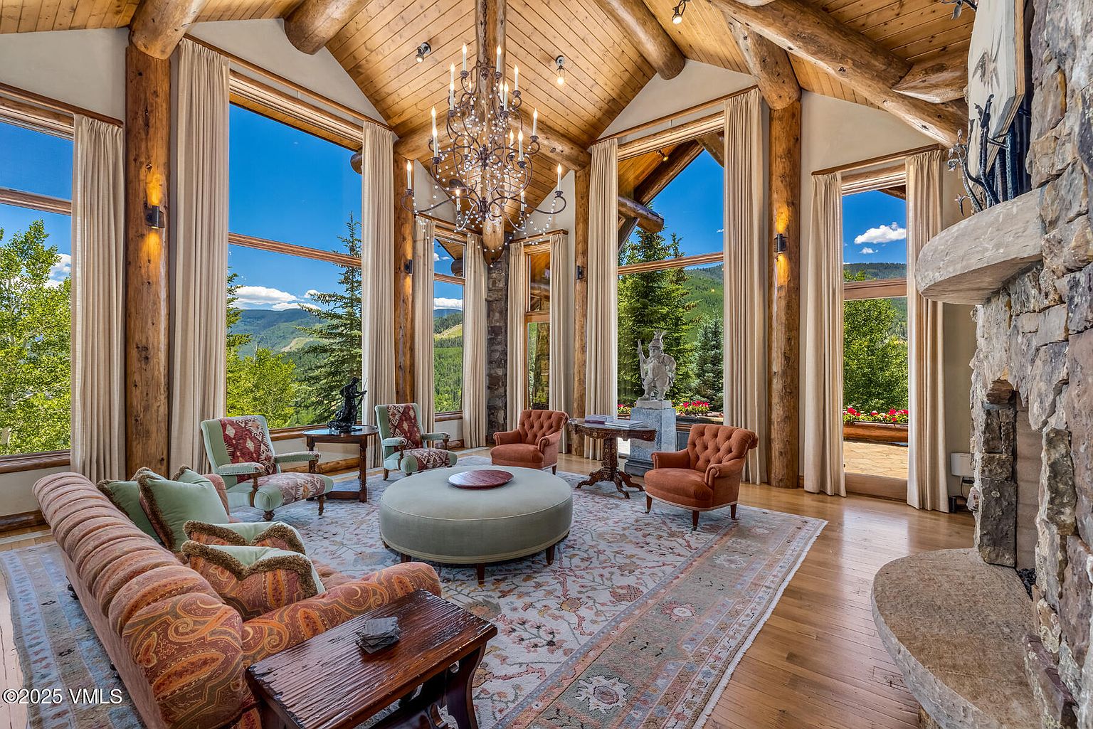 This is a spacious living room featuring a high vaulted ceiling with exposed wooden beams and large windows offering scenic mountain views. The room is furnished with a comfortable sofa, armchairs, and a round ottoman, all arranged on an ornate area rug. A stone fireplace adds a rustic touch, and a chandelier provides elegant lighting, creating a warm and inviting atmosphere.