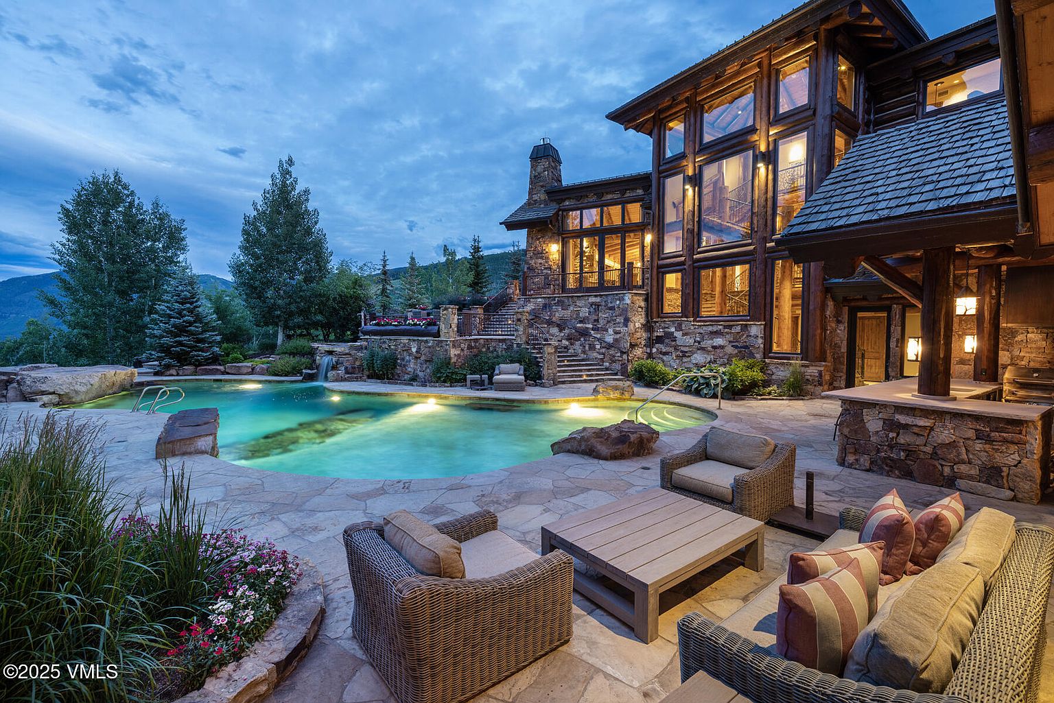 This image showcases a luxurious outdoor pool and spa area of a grand estate. The pool is surrounded by a stone patio with comfortable seating arrangements, including wicker chairs and a sofa with striped pillows. The impressive stone and wood exterior of the house rises in the background, featuring large windows that offer views into the home, creating an inviting and upscale atmosphere.