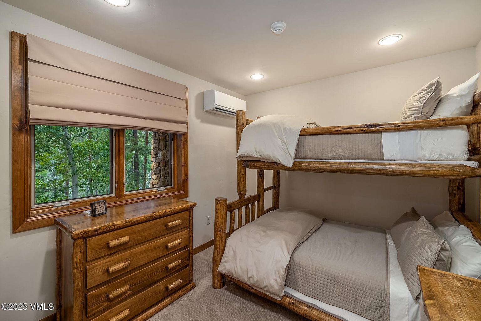 This is a cozy guest bedroom featuring a rustic wooden bunk bed with neutral-toned bedding. A wooden dresser sits beneath a window with a beige roman shade, offering a view of the surrounding trees. The room is well-lit with recessed lighting, creating a warm and inviting atmosphere.