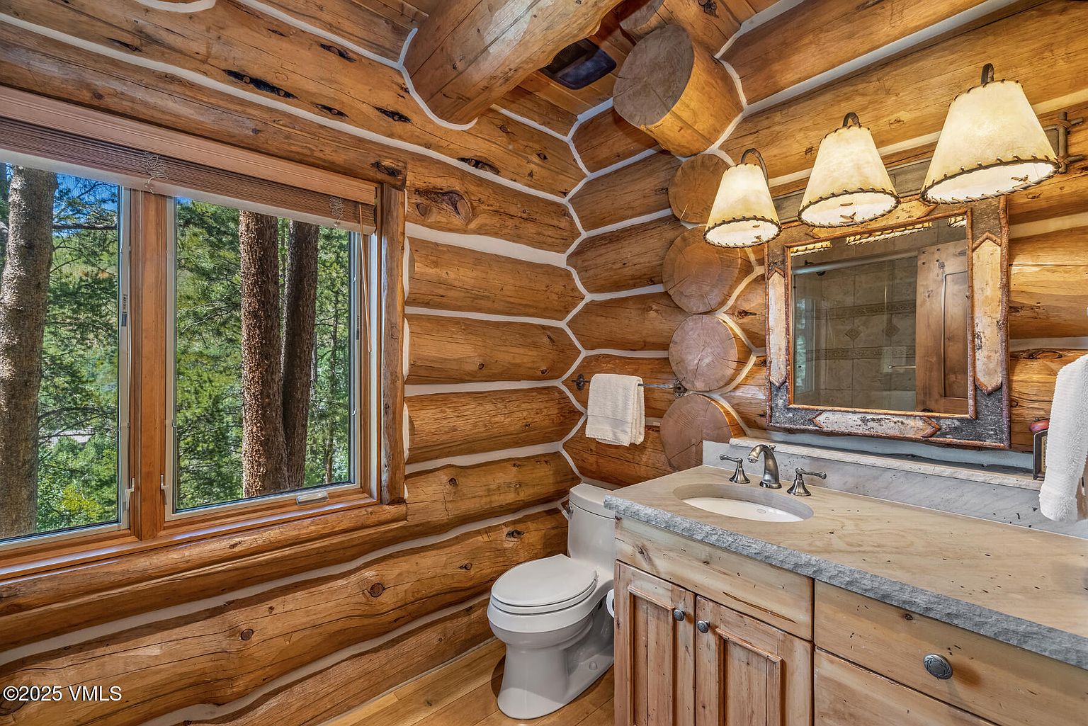 This is a rustic bathroom featuring log walls and ceiling, creating a cozy cabin-like atmosphere. The vanity has a stone countertop and wooden cabinets, complemented by a framed mirror and unique light fixtures. A window provides natural light and a view of the trees outside, enhancing the connection with nature.