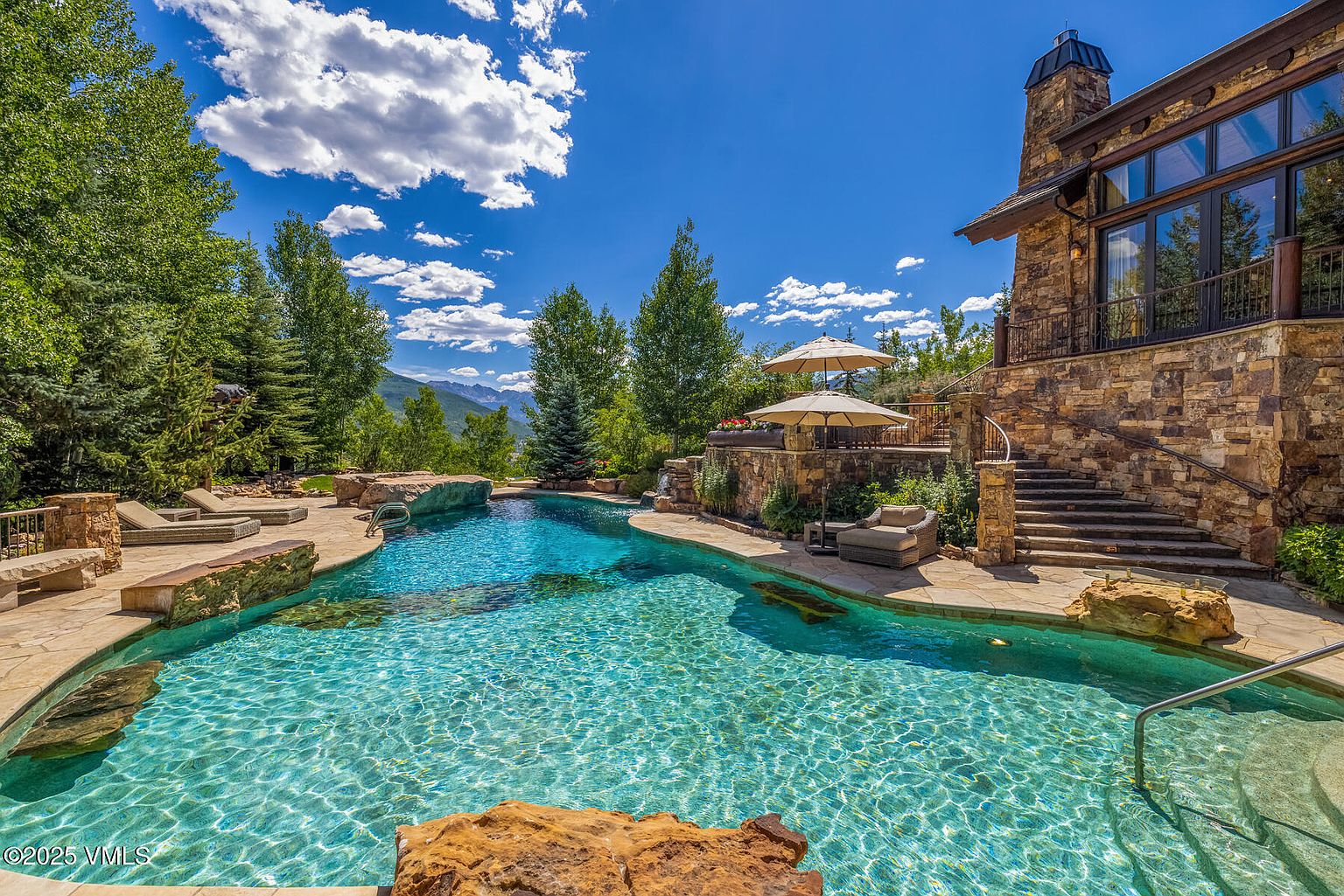 This image showcases a luxurious outdoor pool and spa area, set against a backdrop of lush greenery and a clear blue sky. The pool features a natural stone surround and crystal-clear water, creating an inviting and serene atmosphere. A stone-clad building with large windows and a chimney stands adjacent to the pool, suggesting a high-end residential property with a focus on outdoor living and relaxation.