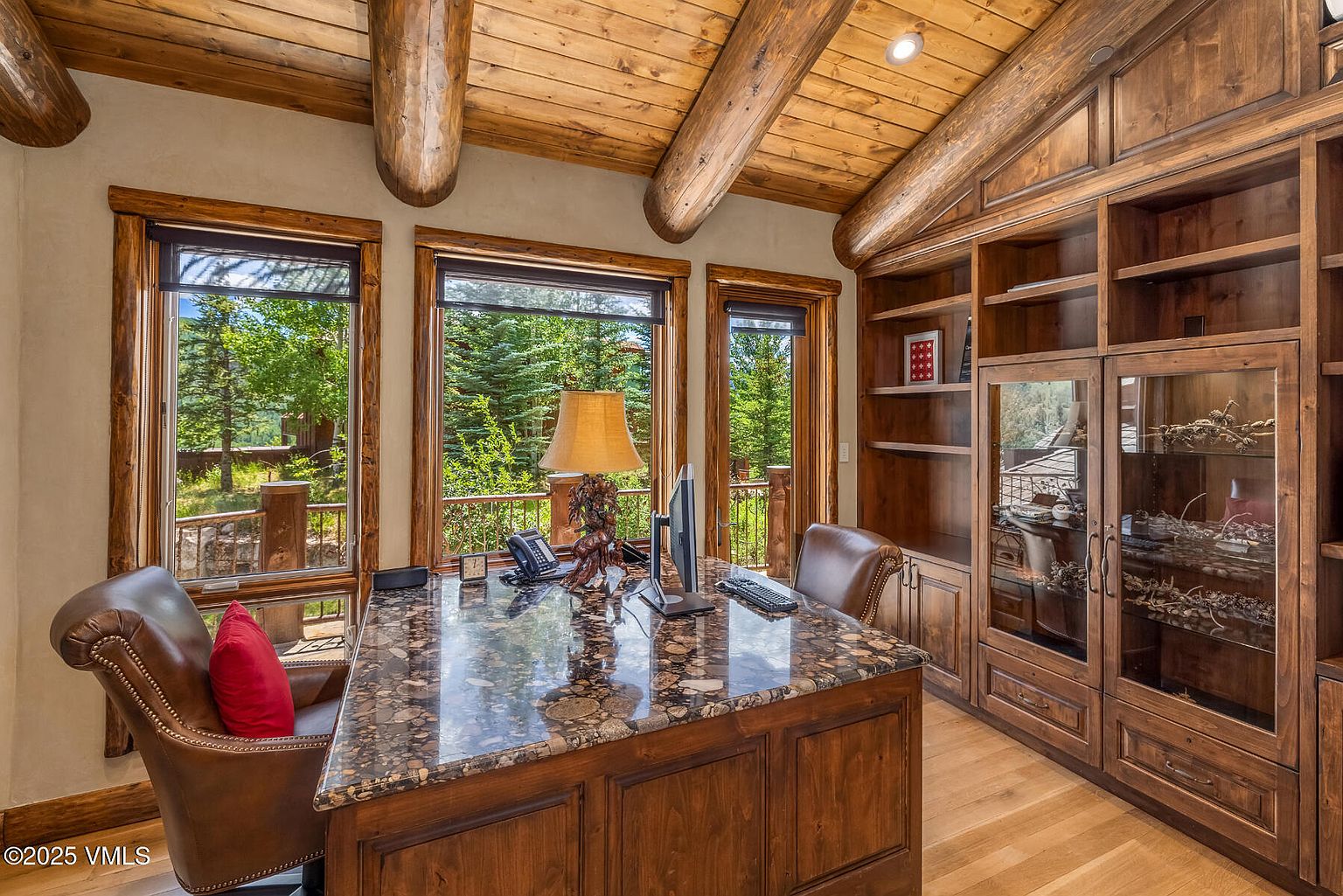 This is an interior shot of a rustic office space featuring a large desk with a granite countertop, surrounded by wooden cabinetry and shelving. The room has a high, wood-beamed ceiling and multiple windows that offer a view of the surrounding trees. The overall impression is one of warmth and sophistication, ideal for a home office.