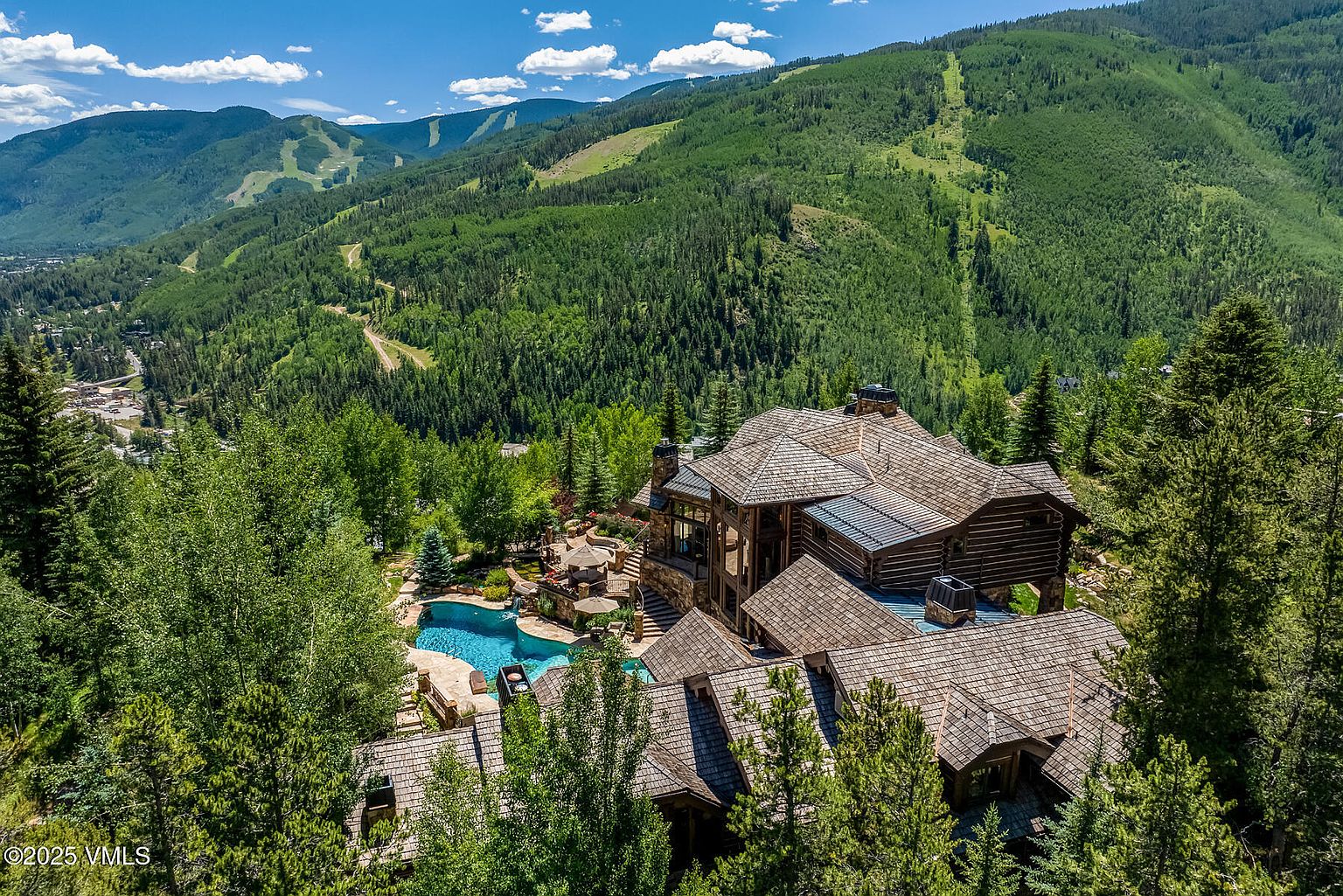 This aerial view showcases a luxurious mountain home with a distinctive wood and stone exterior, complemented by a private pool and meticulously landscaped grounds. The home is nestled amidst lush greenery, with a backdrop of scenic mountains and ski slopes, creating a sense of seclusion and natural beauty. The architectural style emphasizes rustic elegance and integration with the surrounding environment.