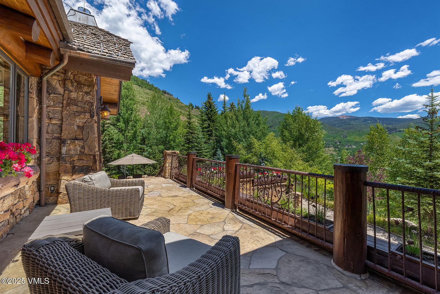 This image showcases a luxurious patio or deck area with comfortable wicker seating and a stone surface. The space is framed by a decorative railing with integrated planters, offering stunning views of the surrounding lush green mountains and a clear blue sky with scattered clouds. The stone and wood elements create a rustic yet elegant ambiance, perfect for outdoor relaxation and entertainment.