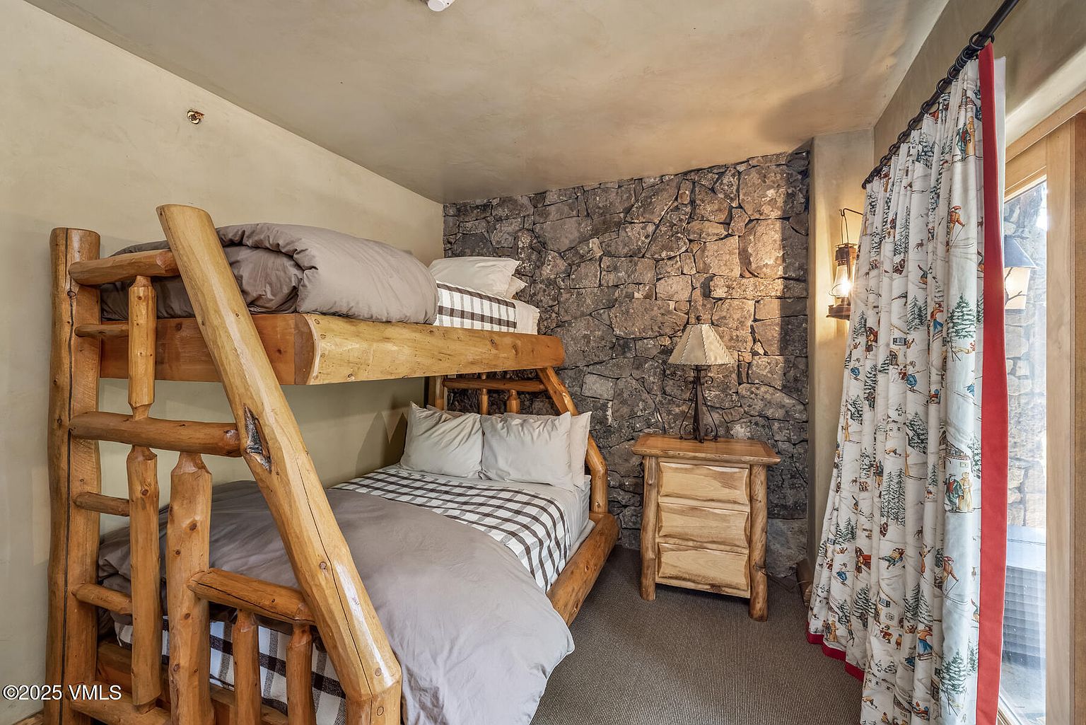 This is a cozy guest bedroom featuring a rustic log bunk bed with gray bedding and a checkered pattern accent. A stone accent wall adds texture, complemented by a wooden nightstand and a lamp. The room is illuminated by a wall-mounted lantern and natural light filtering through a window with patterned curtains, creating a warm and inviting atmosphere.