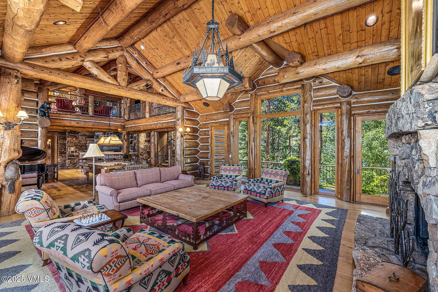 This is a spacious living room with a rustic, log cabin aesthetic. The room features exposed wooden beams, a stone fireplace, and large windows offering natural light and views of the surrounding greenery. The furniture includes patterned armchairs, a sofa, and a large wooden coffee table, all arranged on a colorful area rug. A grand piano is visible in the background, adding to the luxurious feel of the space.