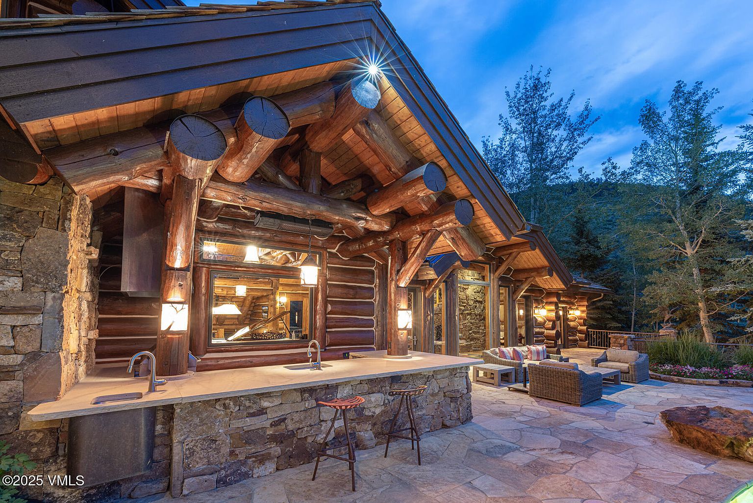 This image showcases a luxurious outdoor patio area featuring a built-in bar with stone accents and wooden stools. Comfortable wicker seating is arranged on a stone patio, with a backdrop of lush greenery and trees. The log cabin architecture adds a rustic yet elegant touch, perfect for entertaining or relaxing outdoors.