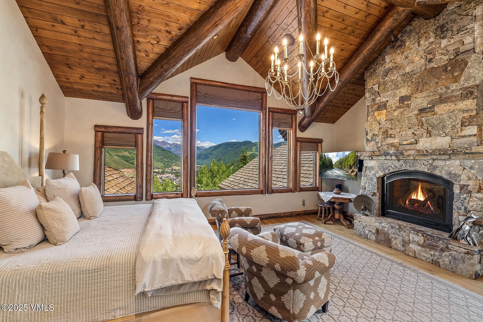 This primary bedroom features a rustic yet luxurious design with exposed wooden beams on the vaulted ceiling and a stone fireplace. Large windows offer scenic mountain views, complemented by comfortable seating and a plush bed. A chandelier adds an elegant touch to the room's warm and inviting atmosphere.