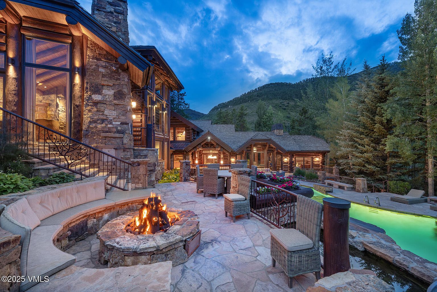 This image showcases a luxurious outdoor patio area featuring a stone fire pit with built-in seating, surrounded by elegant wicker furniture. The patio is part of a grand log and stone home, with a swimming pool visible in the background. The scene is set against a backdrop of lush greenery and a twilight sky, creating a warm and inviting atmosphere.